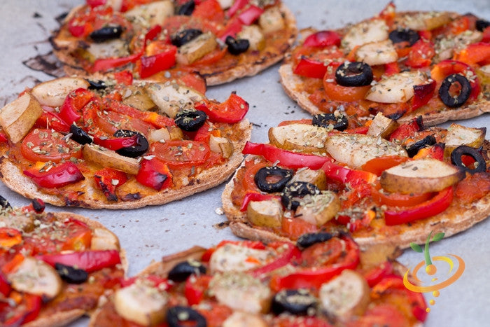 Mini pizzas topped with red pear tomato slices, red bell peppers, black olives, and herbs on a baking tray